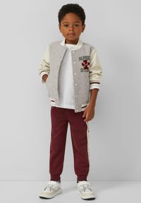 Graue und cremefarbene Bomberjacke mit bestickten Details, kombiniert mit bordeauxfarbenen Jogginghosen mit seitlichen Streifen und weißen Sneakers.