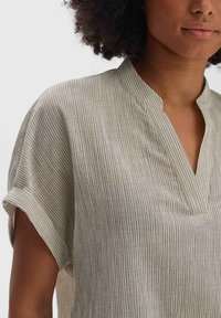 Camicia beige chiaro con strisce verticali, maniche corte e colletto a V. Realizzata in tessuto morbido e testurizzato. Design semplice, senza decorazioni.