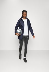 Giacca navale con cappuccio sopra una camicia bianca, pantaloni sportivi grigi con strisce laterali, mentre tiene un pallone da calcio grigio e bianco. Scarpe sportive nere.