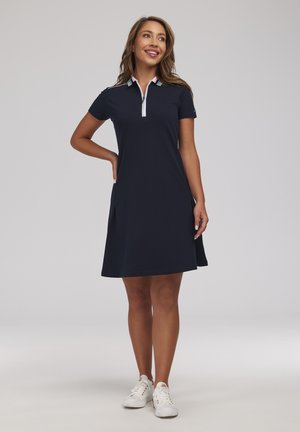 Kobieta uśmiechająca się, ubrana w granatową sukienkę polo z krótkim rękawem i bocznymi rozcięciami oraz białe trampki, stoi z jedną ręką na biodrze.