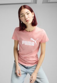 Rožinė medvilninė marškinė su balta "PUMA" logotipo ir silueto dizainu. Paprastas apvalus iškirpimas, trumpomis rankovėmis ir minkšta tekstūra. Suderinta su šviesiai mėlynomis džinsomis.