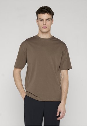 Man with curly dark hair wearing a plain brown short-sleeve T-shirt and dark pants, standing against a light gray background.