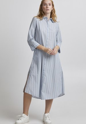 Vestito camicia a righe azzurro chiaro, con bottoni frontali, maniche 3/4, lunghezza ginocchio con spacchi laterali, abbinato a sneakers bianche. Tessuto in cotone morbido.