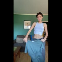 Valkoinen crop-top, harmaat housut, pitämässä vihreää paitaa. Sisätilat, jossa vihreä seinä, sohva ja kehystetty taide. Puulattia ja pöytä näkyvissä.