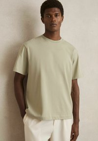 Tricou din bumbac de culoare verde deschis, cu mâneci scurte, guler rotund și croială lejeră, purtat cu pantaloni de culoare deschisă pe un fundal neutru.