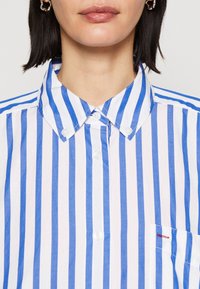 Camisa a rayas con rayas verticales alternas en azul y blanco, cuello abotonado, y un pequeño bolsillo en el pecho con costura de acento roja.