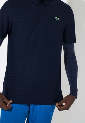 Mann trägt ein marineblaues Polohemd mit einem kleinen grünen Krokodil-Logo und hellblauen Hosen, hält den Saum des Hemdes an beiden Seiten.