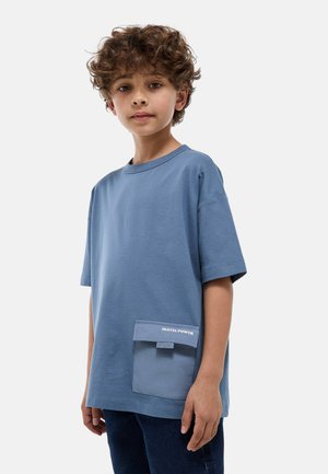 Garçon aux cheveux bouclés portant un t-shirt bleu ample avec une poche avant étiquetée "PASTEL POWER" et un pantalon foncé sur un fond uni.