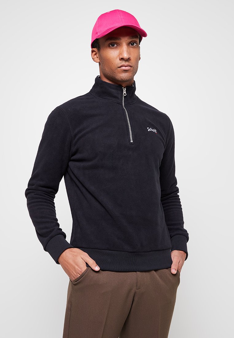 Maglione in pile nero con collo a mezza zip e polsini a coste. Presenta un piccolo logo sul petto. Abbinato a un cappellino rosa acceso.