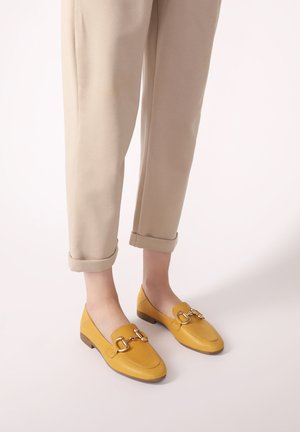 Gelbe Lederloafers mit flacher Sohle, versehen mit einem goldenen Kettendetail auf dem Vorderblatt, getragen mit hochgerollten beigen Hosen.