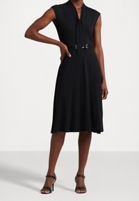 Černé šaty bez rukávů s vázáním u krku, přiléhavým pasem a stříbrnými přezkami. Hladká látka s lehce splývavou sukní do A.