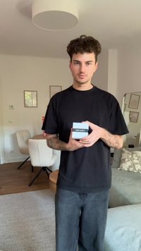 Black t-shirt and dark jeans worn by a person holding a gray Calvin Klein box. Light interior with furnishings visible in the background.