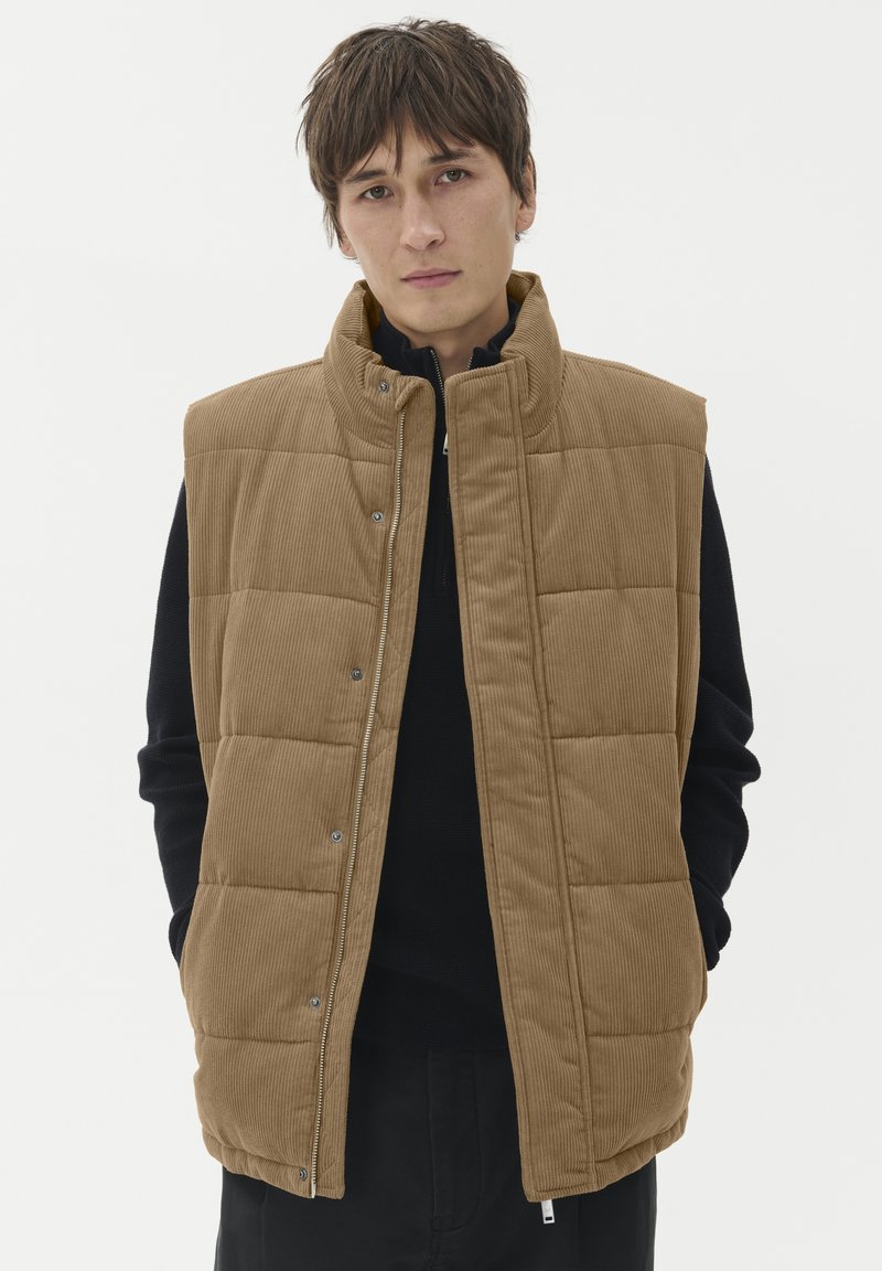 Braun gestreifter Weste mit Reißverschluss und Druckknopfverschluss, ausgestattet mit gestepptem Design und hohem Kragen, getragen über einem schwarzen Pullover.