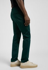 Chinos verdi in misto cotone, con vestibilità affusolata, tasca posteriore e abbinati a sneakers di colore chiaro. Texture liscia.
