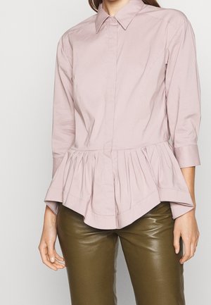 Vrouw draagt een lichtroze blouse met lange mouwen, een spitse kraag en een geplooide peplumzoom, gecombineerd met olijfgroene leren broek.