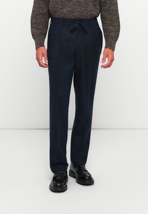 Man wearing dark navy tailored trousers with front crease, black leather loafers, and a brown patterned knit sweater.