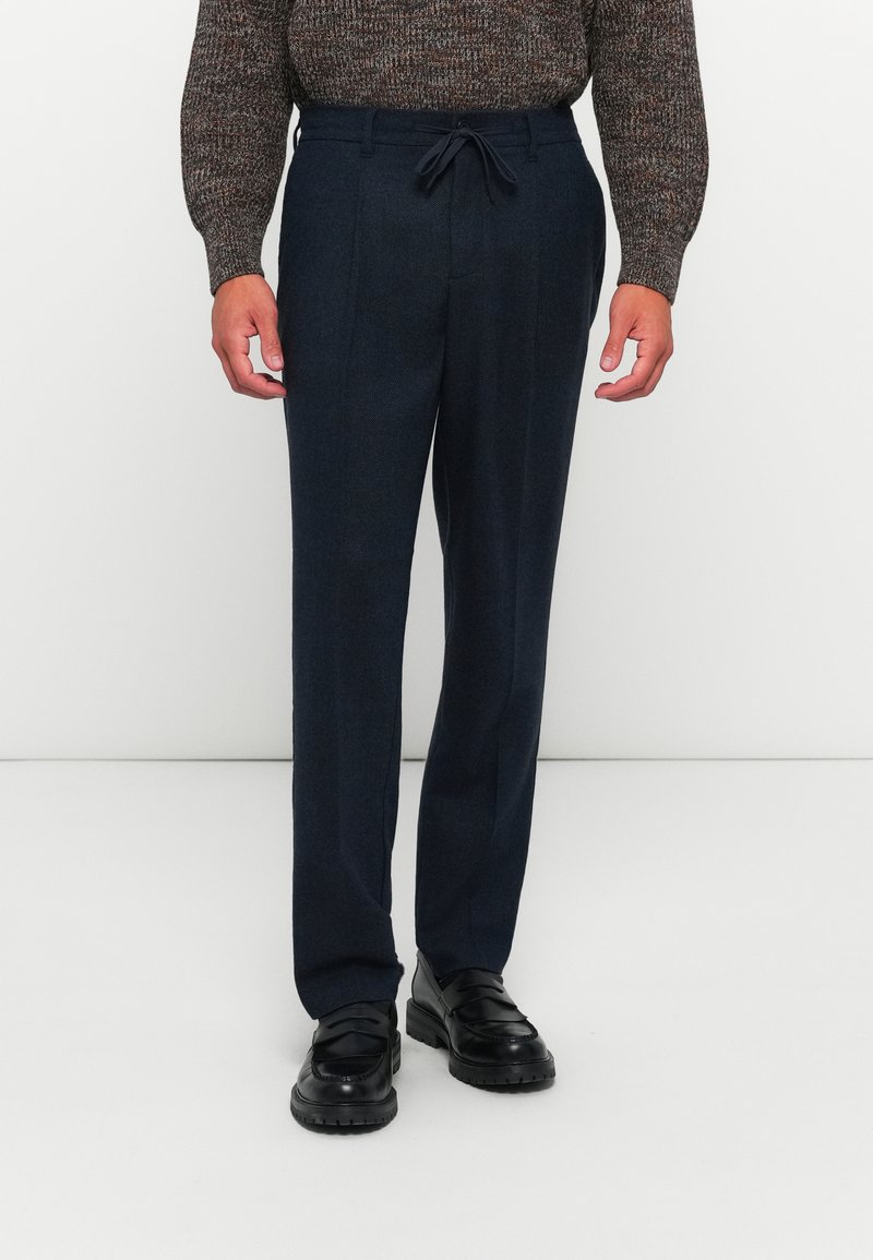 Man in een donkerblauwe op maat gemaakte broek met een vorenplooien, zwarte leren instappers en een bruine gebreide trui met patroon.