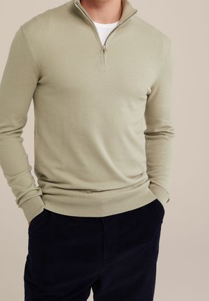 Mann in einem hellbeigen Pullover mit halbem Reißverschluss über einem weißen Unterhemd, mit dunkeln marineblauen Hosen, die Hände in den Taschen, vor neutralem Hintergrund.