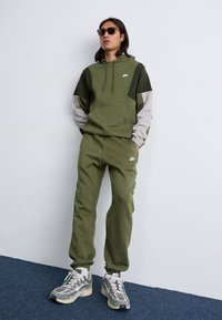 Olívazöld Nike melegítőnadrágban, kapucnival és szürke ujjakkal rendelkező személy napszemüveget viselve, egy sima fal és kék szőnyeg előtt áll.