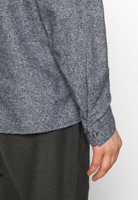 Szara, teksturowana koszula z długim rękawem i mankietem zapinanym na guziki, zestawiona z ciemnym materiałem o subtelnym wzorze poniżej.