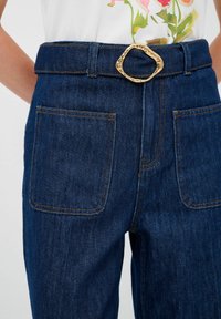 Jeans in denim blu scuro con una cintura ampia e una fibbia a forma ovale in oro testurizzato. Presentano due tasche posteriori con cuciture a contrasto.