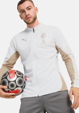 Uomo che indossa una giacca da calcio Puma bianca e beige, che tiene in mano un pallone da calcio nero e rosso con motivo su sfondo semplice.