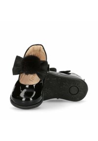 Scarpe in vernice nera con punta rotonda, caratterizzate da un fiocco nero testurizzato e un accento a pom-pom. La suola è decorata con un motivo per migliorare la trazione.
