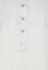 Blusa grigia chiara con una patta bottonata caratterizzata da sei bottoni argentati; tessuto liscio e leggermente lucido; design con colletto.