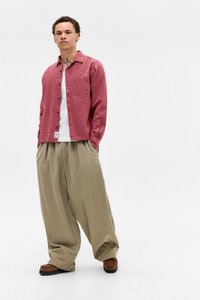 Rotes kariertes Hemd, weißes T-Shirt darunter, weite beige Cordhose, braune Schuhe, steht vor einem weißen Hintergrund.