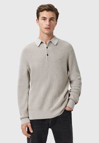 Lichtgrijze, geribbelde polo met lange mouwen en een witte kraag, drie zwarte knopen en een klein embleem op de borst. Draag het met een donkere jeans.