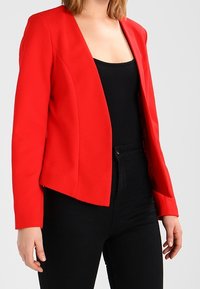 Červený blazer se strukturovaným designem, přiléhavým siluetou a otevřeným předním dílem. Vyrobeno z texturované látky, kombinováno s černým topem a tmavými džíny.