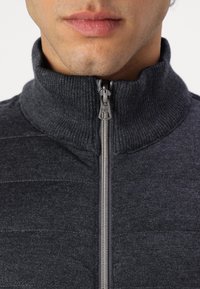 Veste zippée grise avec un col haut, présentant une texture côtelée et une fermeture éclair en métal. Une couture horizontale apporte un intérêt visuel.