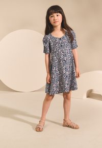 Lyhythihainen tummansininen mekko, jossa on vaaleanpunainen kukkakuosi, A-linjainen malli, pyöreä pääntie ja pussihihat. Yhdistetty vaaleanpunaisiin remmisandaaleihin.