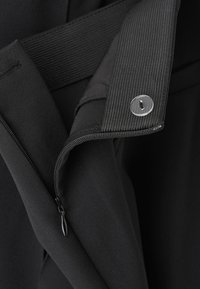 Černý textilní oděv s kapsou na zip, texturovanými akcenty a detailním patentním knoflíkem. Hladký a strukturovaný materiál.