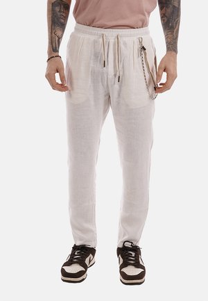 Uomo che indossa pantaloni di lino bianchi con coulisse, un accessorio a catena e scarpe da ginnastica bianche e nere, in piedi con le braccia tatuate visibili.