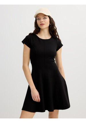 Mujer con cabello largo y ondulado, que lleva un vestido corto negro y una gorra de béisbol beige, de pie frente a un fondo claro liso.