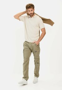 Beżowa koszulka polo, oliwkowe spodnie cargo z bocznymi kieszeniami, białe sneakersy i brązowy sweter narzucony na ramię.