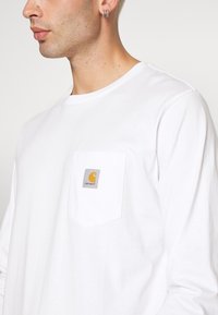 Langermet hvit bomullsskjorte med brystlomme og et firkantet Carhartt-merke i grått med gult logo. Glatt tekstur, rund hals-design.