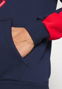 Marinblå sweatshirt med röda accenter, featuring en känguruficka, ribbade muddar och en texturerad yta. Synlig hand som vilar i fickan.