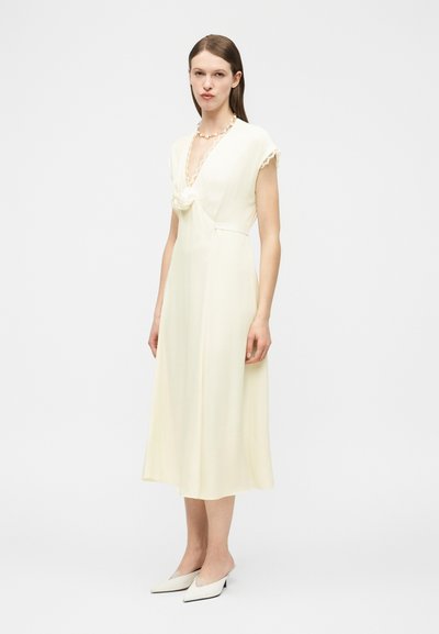 Femme portant une robe midi couleur crème avec des manches courtes, un décolleté en V profond, un détail floral au niveau de la poitrine, et des chaussures blanches pointues.