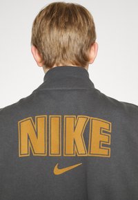 Donkergrijze sweatshirt met de tekst "NIKE" in grote geel-oranje letters op de achterkant, met daaronder het swoosh-logo in dezelfde kleur.