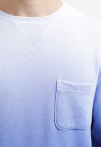 Sudadera de cuello redondo azul claro con un pequeño bolsillo en el pecho y detalle cosido debajo del cuello, llevada por una persona.