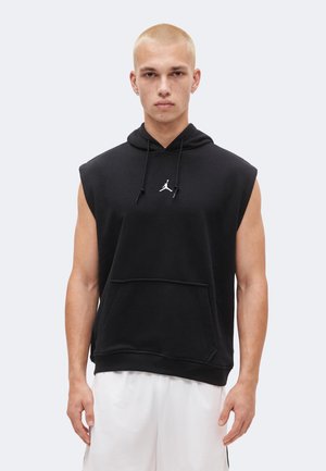 Νέος άνδρας με κοντά ξανθά μαλλιά, φοράει μαύρο αμάνικο φούτερ με λευκό λογότυπο και λευκά αθλητικά σορτς, κοιτάζοντας μπροστά με απλό φόντο.