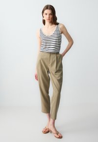 Gestreept tanktop in wit en marine; beige, op maat gemaakte broeken met een relaxte pasvorm. Model draagt tan gekleurde haksandalen en staat tegen een neutrale achtergrond.