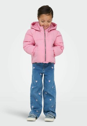 Junges Mädchen, das lächelt und nach unten schaut, trägt eine pinke Kapuzenjacke und blaue Jeans mit pinken Herzmustern.