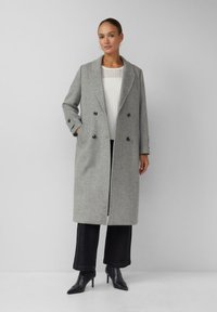 Manteau gris en laine à double boutonnage avec un col à revers cranté, des poches latérales et des détails de boutons, associé à un haut blanc côtelé et un pantalon noir à jambes larges.