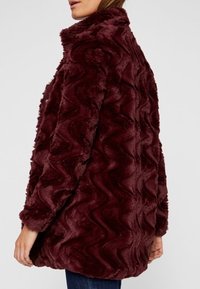 Bordowy płaszcz z sztucznego futra o teksturze zygzaków, z wysokim kołnierzem i luźnym krojem, widziany z tyłu. Miękki i puszysty materiał.