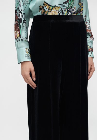 Personne portant une jupe en velours noire et une chemise à manches longues bleu clair imprimée d’illustrations colorées de chevaux et de carrosses.
