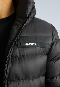 Fekete puff jacket, magas gallérral, vízszintes steppelt részekkel és fekete szövetcímkével, rajta fehér betűkkel a "JACK1T" felirattal.