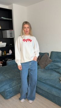 Ξανθιά γυναίκα που στέκεται στο σαλόνι, φορώντας λευκό φούτερ με κόκκινες καρδιές και τη λέξη "HUGO", μπλε τζιν με φαρδιά πόδια και λευκά αθλητικά παπούτσια.